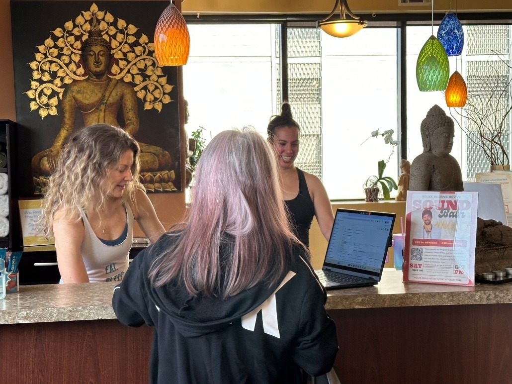 Two smiling yoga teachers greet a student as she checks in at the front desk. There is a large painting of Buddha in the background next to 3 large windows.